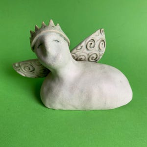 May include: A white ceramic sculpture of a winged figure wearing a crown. The figure has its eyes closed and is lying on its side. The wings are detailed with swirling patterns.