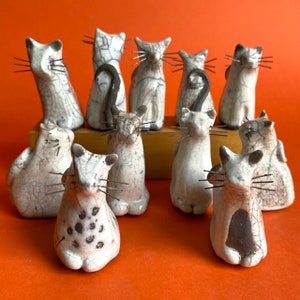 May include: A collection of 12 ceramic cat figurines, each with a unique crackled glaze finish. The cats are in various poses, some sitting, some standing, and some lying down. They are all white with black accents.