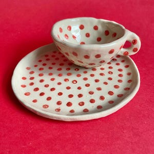 May include: A handmade ceramic teacup and saucer set. The off-white pieces are decorated with red polka dots. The cup has a handle and sits on a matching saucer. The set is on a red background.