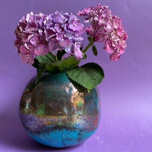 May include: A decorative vase with a metallic finish, holding vibrant purple and pink hydrangea flowers. The vase has a rounded shape with a teal and bronze colour scheme. The background is a solid purple.