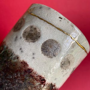 May include: A ceramic cup featuring a light grey crackle glaze finish with dark grey circular spots. The upper rim is repaired with a gold-coloured line. The lower section of the cup transitions into a dark, textured, mottled brown and black surface.