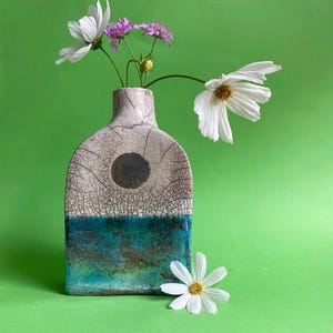 May include: A handmade ceramic vase with a distinctive crackle glaze. The vase has a dark blue circle and a turquoise lower section. White cosmos flowers and purple scabiosa are arranged inside the vase, against a green backdrop.