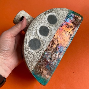May include: A ceramic vase with an unusual, asymmetrical shape. It has a white, cracked glaze on one side with three grey circles. The other side has an iridescent, colourful finish with orange, blue, and green hues. A small handle extends from the top.