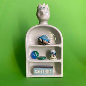 May include: White ceramic shelf unit with three shelves. The top shelf features a ceramic turtle figurine. The middle shelf has two round buttons, one blue and one green. The bottom shelf has a small box with the word "CAFETERIA" printed on it.