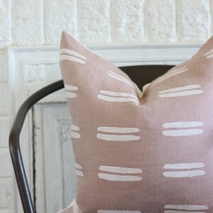 May include: A blush-colored decorative pillow with a repeating pattern of white horizontal stripes. The pillow is placed on a dark brown metal chair, offering a touch of modern design to any living space.
