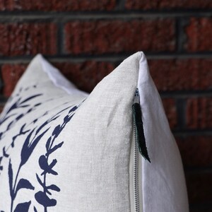 Pu&ograve; includere: Un cuscino decorativo con una fodera in lino bianco con una stampa botanica blu navy. Il cuscino ha una chiusura a cerniera nascosta con una linguetta verde scuro. Lo sfondo &egrave; un muro di mattoni rossi.