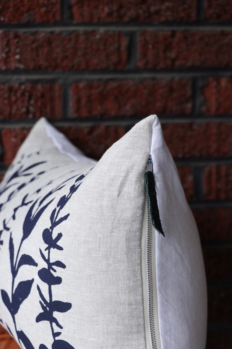 Pu&ograve; includere: Un cuscino decorativo con una fodera in lino bianco con una stampa botanica blu navy. Il cuscino ha una chiusura a cerniera nascosta con una linguetta verde scuro. Lo sfondo &egrave; un muro di mattoni rossi.