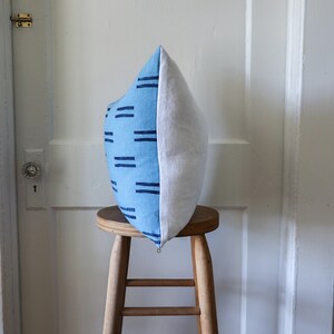 Puede incluir: Una almohada con estampado azul y blanco se encuentra sobre un taburete de madera. La almohada tiene un patr&oacute;n geom&eacute;trico de l&iacute;neas azules sobre un fondo azul claro.