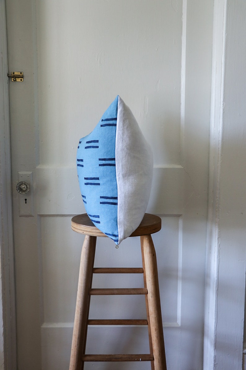 Puede incluir: Una almohada con estampado azul y blanco se encuentra sobre un taburete de madera. La almohada tiene un patr&oacute;n geom&eacute;trico de l&iacute;neas azules sobre un fondo azul claro.