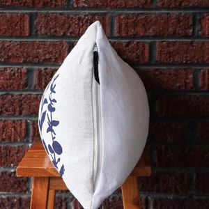 Pu&ograve; includere: Cuscino decorativo bianco con un motivo floreale blu navy su un lato. Il cuscino ha una chiusura a cerniera ed &egrave; posto su uno sgabello di legno. Lo sfondo &egrave; un muro di mattoni rossi.