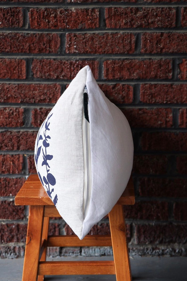 Pu&ograve; includere: Cuscino decorativo bianco con un motivo floreale blu navy su un lato. Il cuscino ha una chiusura a cerniera ed &egrave; posto su uno sgabello di legno. Lo sfondo &egrave; un muro di mattoni rossi.