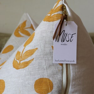 Puede incluir: Dos cojines decorativos de lino color crema con un estampado floral amarillo mostaza. Los cojines tienen una cremallera de lat&oacute;n y una etiqueta que dice "MUSE textiles hand printed home goods".