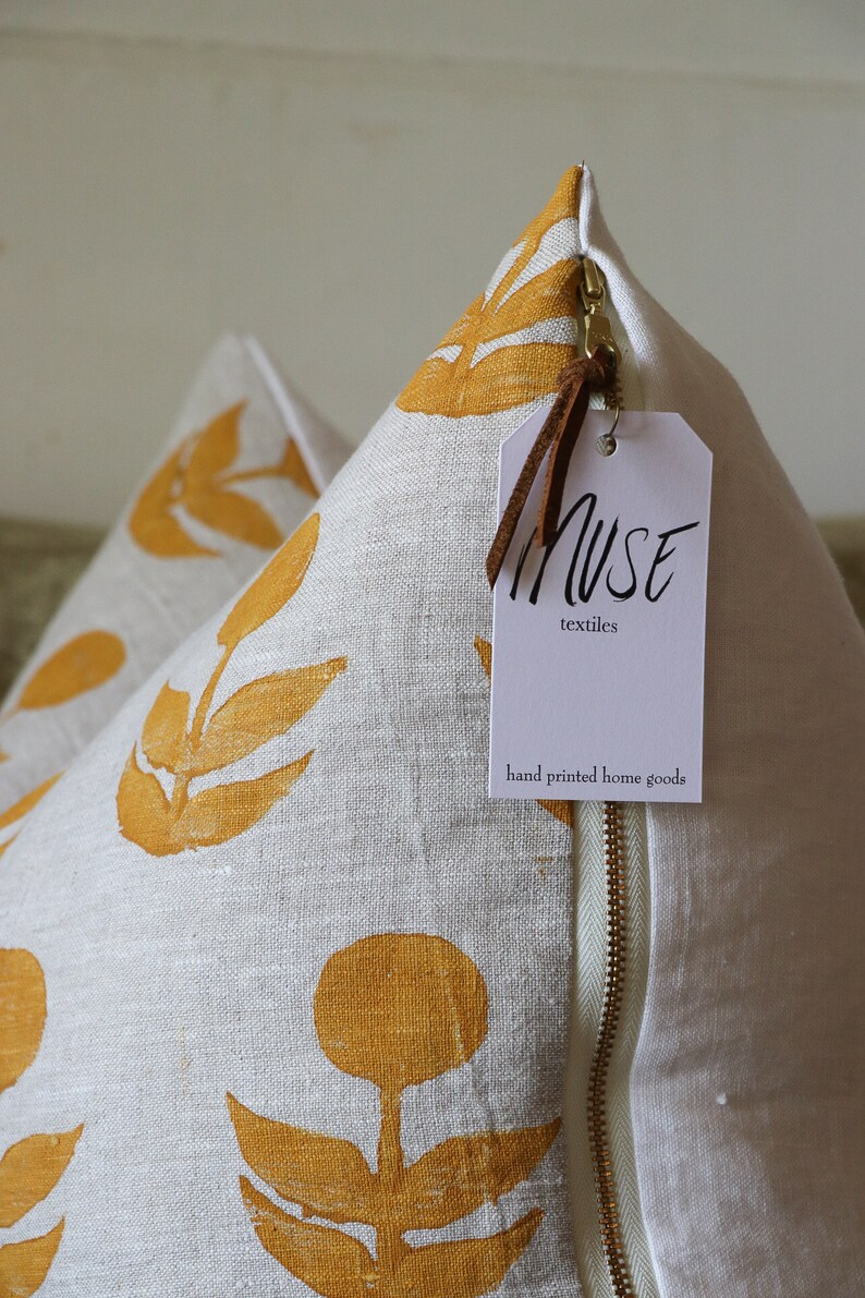Puede incluir: Dos cojines decorativos de lino color crema con un estampado floral amarillo mostaza. Los cojines tienen una cremallera de lat&oacute;n y una etiqueta que dice "MUSE textiles hand printed home goods".