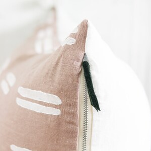 May include: Close-up of a decorative pillow with a two-tone design. One side is a dusty rose color with white abstract shapes, and the other is white. A silver zipper and a dark green pull are visible.