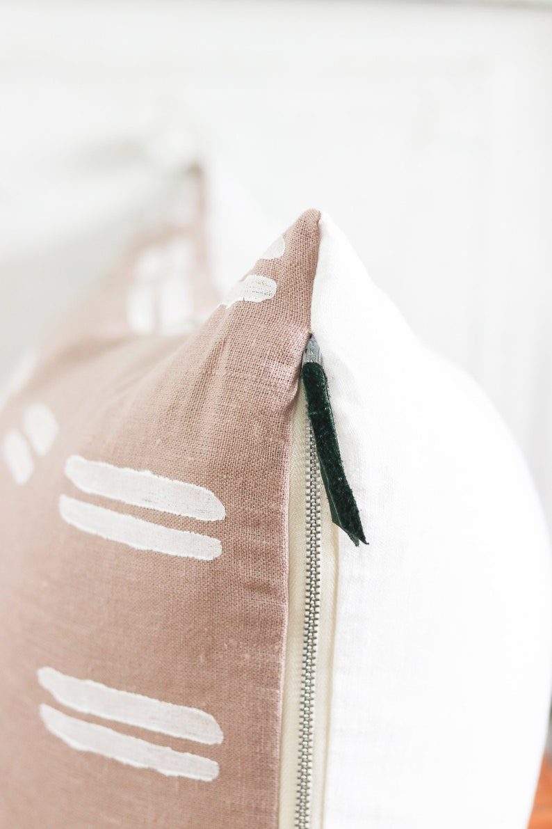 May include: Close-up of a decorative pillow with a two-tone design. One side is a dusty rose color with white abstract shapes, and the other is white. A silver zipper and a dark green pull are visible.