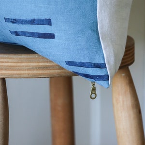 Puede incluir: Una almohada de lino azul con la parte posterior blanca y cierre de cremallera. La almohada tiene un patr&oacute;n geom&eacute;trico de l&iacute;neas azules oscuras.