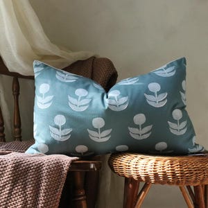 May include: A rectangular teal pillow with a white floral pattern rests on a wooden chair and a wicker stool. The pillow's design features repeating stylized flower shapes. A brown knitted throw is draped over the chair.
