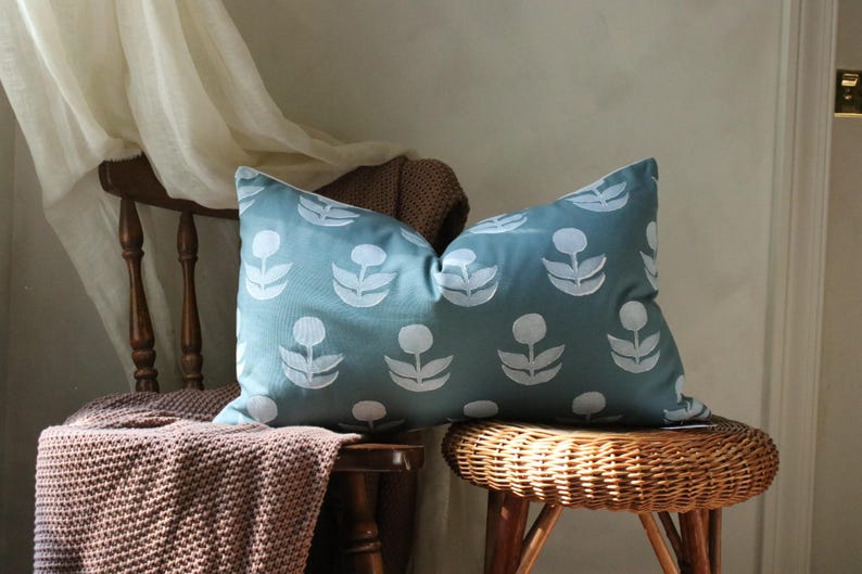May include: A rectangular teal pillow with a white floral pattern rests on a wooden chair and a wicker stool. The pillow's design features repeating stylized flower shapes. A brown knitted throw is draped over the chair.