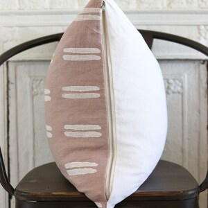 May include: A decorative pillow with a two-tone design. One side features a dusty rose fabric with white horizontal stripe patterns. The other side is plain white. A zipper runs along the edge. The pillow is on a dark metal chair.