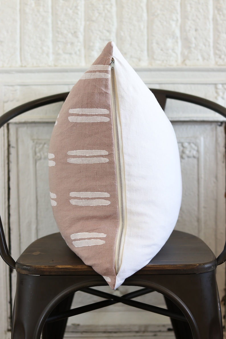 May include: A decorative pillow with a two-tone design. One side features a dusty rose fabric with white horizontal stripe patterns. The other side is plain white. A zipper runs along the edge. The pillow is on a dark metal chair.