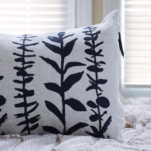 Pu&ograve; includere: Cuscino decorativo rettangolare con sfondo grigio chiaro e motivo ripetuto di illustrazioni botaniche blu navy scuro. Il cuscino &egrave; realizzato in tessuto strutturato ed &egrave; posto su una coperta strutturata vicino a una finestra.