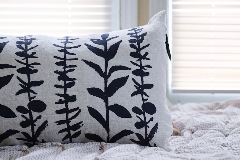 Pu&ograve; includere: Cuscino decorativo rettangolare con sfondo grigio chiaro e motivo ripetuto di illustrazioni botaniche blu navy scuro. Il cuscino &egrave; realizzato in tessuto strutturato ed &egrave; posto su una coperta strutturata vicino a una finestra.