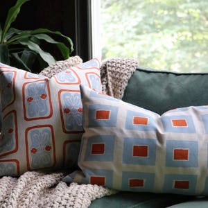 May include: Two decorative throw pillows featuring geometric patterns. One square pillow displays a light blue floral motif within orange rectangular borders, while the rectangular pillow shows a grid of light blue squares with orange centers.