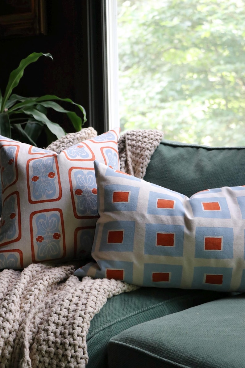 May include: Two decorative throw pillows featuring geometric patterns. One square pillow displays a light blue floral motif within orange rectangular borders, while the rectangular pillow shows a grid of light blue squares with orange centers.