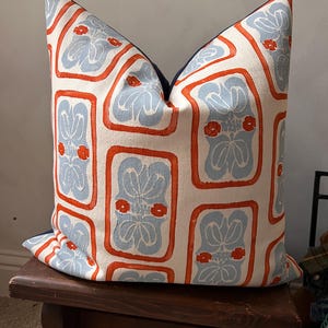May include: Decorative throw pillow with a repeating pattern of light blue floral designs within orange rectangular frames on an off-white background. The pillow has a dark blue border.