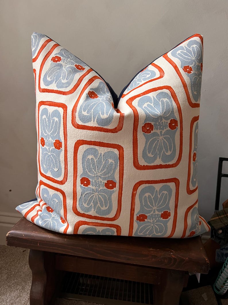 May include: Decorative throw pillow with a repeating pattern of light blue floral designs within orange rectangular frames on an off-white background. The pillow has a dark blue border.