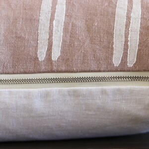 May include: Close-up of a decorative pillow with a two-tone design. The top section is a dusty rose color with vertical white brushstroke patterns. The bottom section is a light cream color, with a silver zipper.