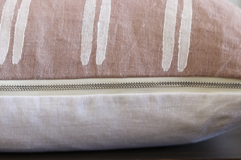 May include: Close-up of a decorative pillow with a two-tone design. The top section is a dusty rose color with vertical white brushstroke patterns. The bottom section is a light cream color, with a silver zipper.