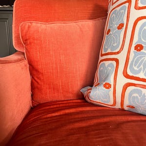 May include: A close-up of a red-orange velvet sofa with two pillows. One pillow is a solid red-orange color, and the other is white with a blue and red floral pattern. The sofa has a high back and armrests.