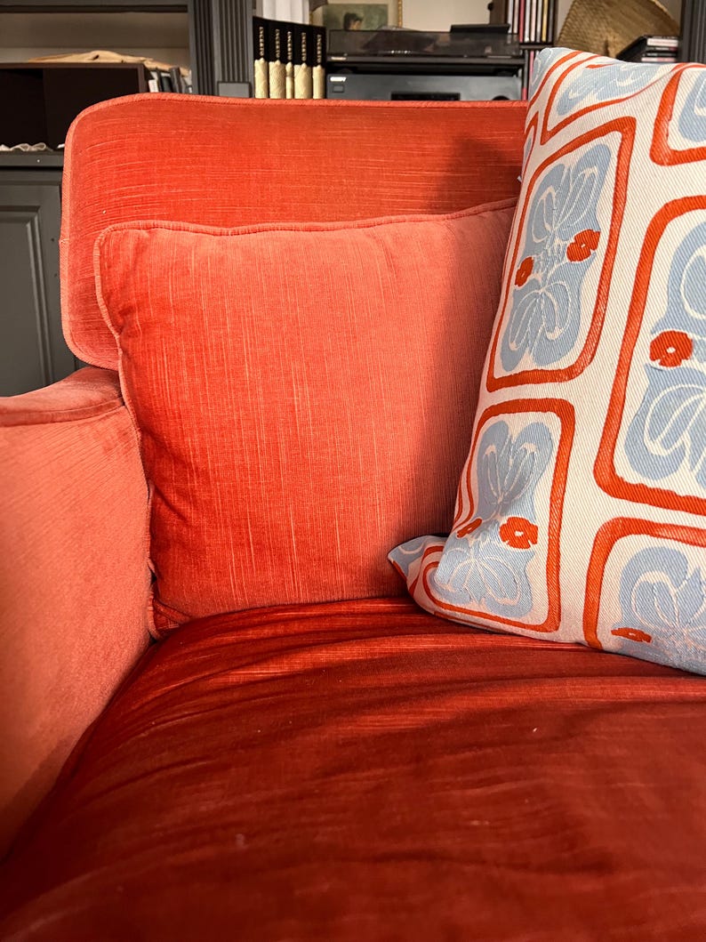 May include: A close-up of a red-orange velvet sofa with two pillows. One pillow is a solid red-orange color, and the other is white with a blue and red floral pattern. The sofa has a high back and armrests.