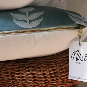 May include: Close-up of a decorative pillow with a teal floral pattern and a gold zipper. The pillow is white on the bottom and rests on a woven basket. A tag reads "MUSE textiles hand printed home goods."