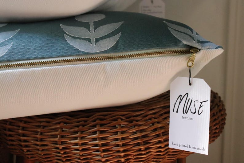 May include: Close-up of a decorative pillow with a teal floral pattern and a gold zipper. The pillow is white on the bottom and rests on a woven basket. A tag reads "MUSE textiles hand printed home goods."