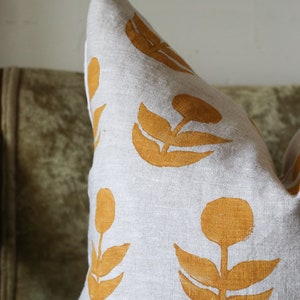 Puede incluir: Un coj&iacute;n decorativo con tela de lino gris claro y un patr&oacute;n repetido de dise&ntilde;os florales de color mostaza. El coj&iacute;n tiene forma cuadrada y suave, perfecto para a&ntilde;adir un toque de estilo a cualquier habitaci&oacute;n.