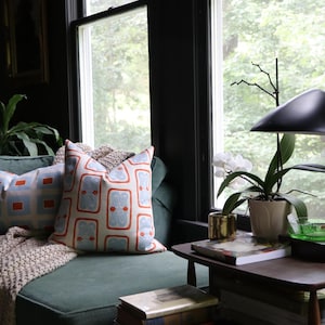 May include: A green sofa with two patterned throw pillows featuring orange and light blue geometric designs, a beige knit throw blanket, a wooden side table, a black desk lamp, a white orchid in a pot, and a small blue glass bird figurine.