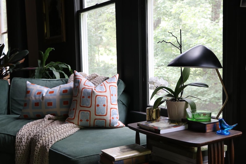 May include: A green sofa with two patterned throw pillows featuring orange and light blue geometric designs, a beige knit throw blanket, a wooden side table, a black desk lamp, a white orchid in a pot, and a small blue glass bird figurine.