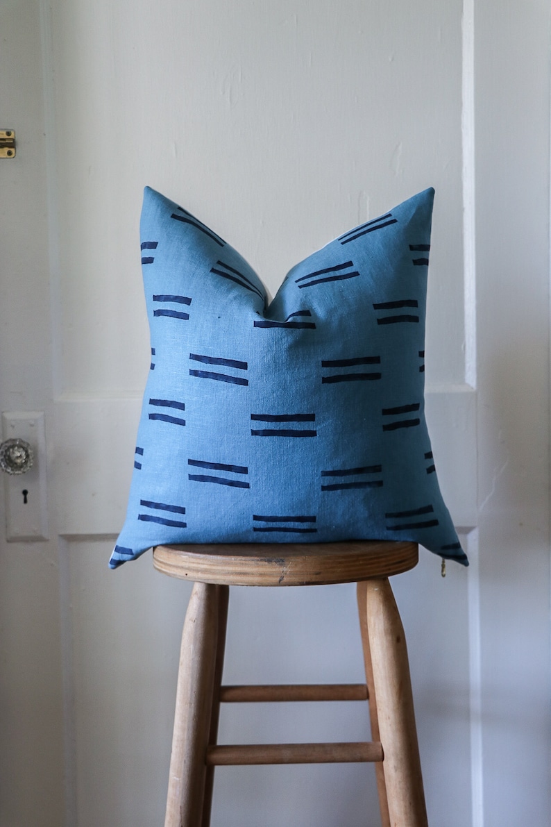 Puede incluir: Una almohada azul con un patr&oacute;n geom&eacute;trico de rayas negras. La almohada est&aacute; sentada en un taburete de madera.