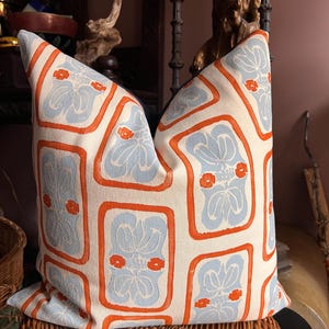 May include: Decorative throw pillow with a repeating pattern of light blue floral designs within orange rectangular frames on an off-white background. The pillow is placed on a woven wicker basket, suggesting a home decor theme.