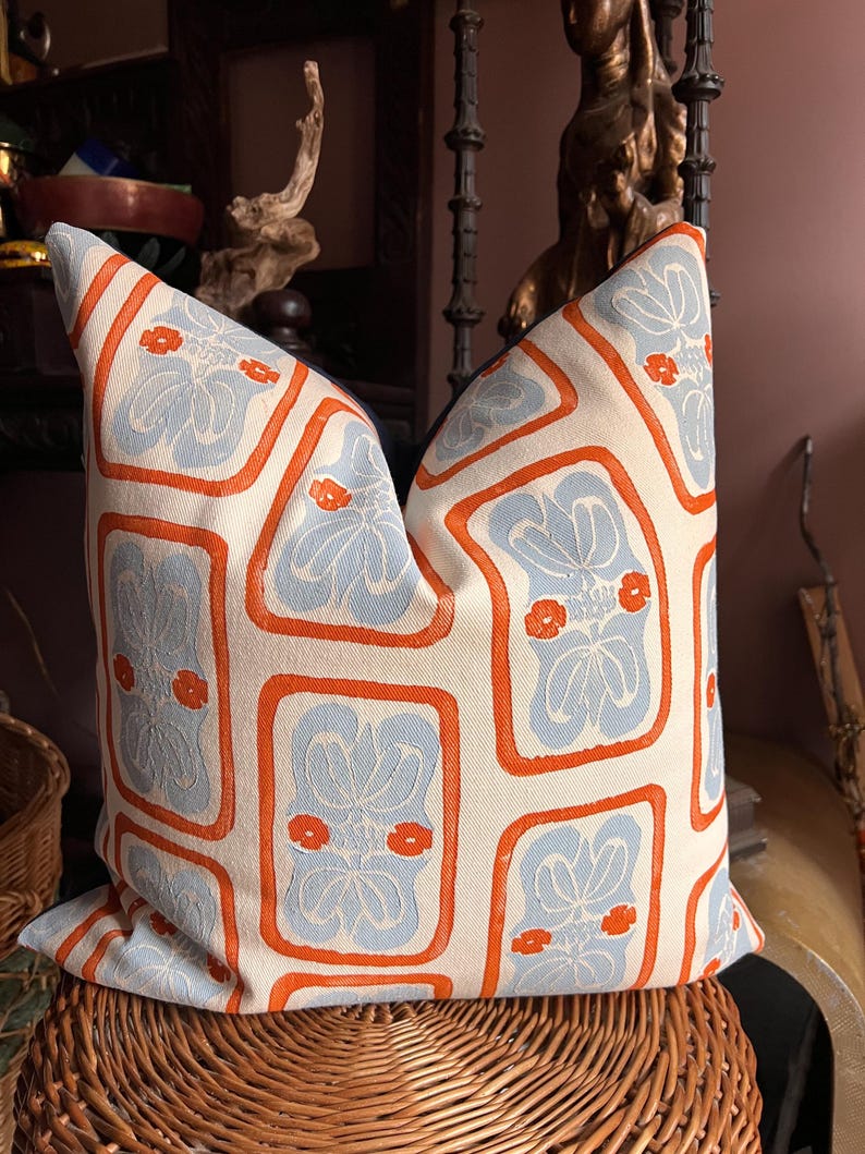 May include: Decorative throw pillow with a repeating pattern of light blue floral designs within orange rectangular frames on an off-white background. The pillow is placed on a woven wicker basket, suggesting a home decor theme.