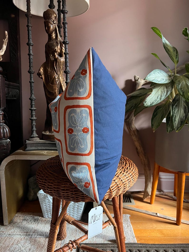 May include: A decorative pillow with a blue and floral patterned design. The pillow has a solid blue side and a patterned side with orange and gray floral motifs. The pillow is displayed on a wicker chair.