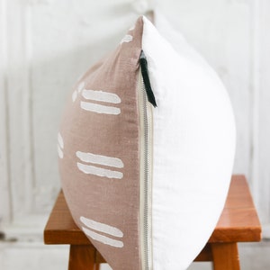 May include: A decorative pillow with a two-tone design. One side is a light brown with white horizontal stripe patterns, and the other side is white. The pillow has a silver zipper and is placed on a wooden stool.