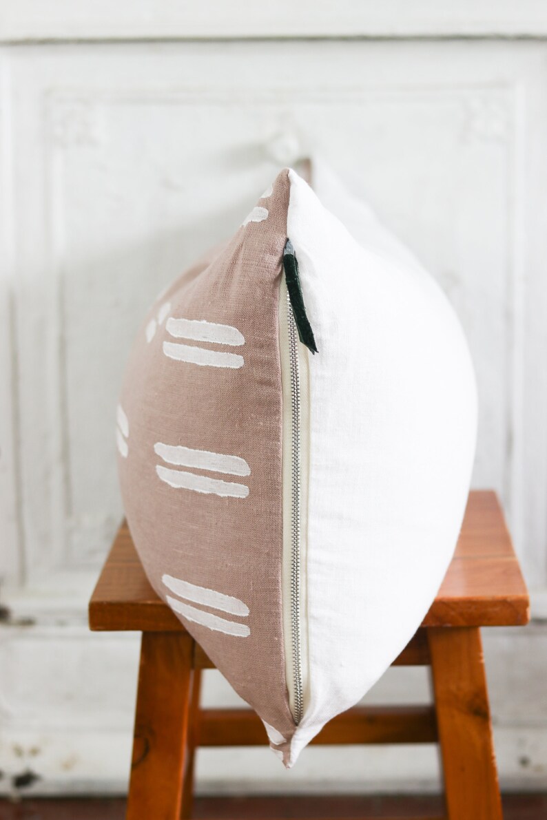 May include: A decorative pillow with a two-tone design. One side is a light brown with white horizontal stripe patterns, and the other side is white. The pillow has a silver zipper and is placed on a wooden stool.