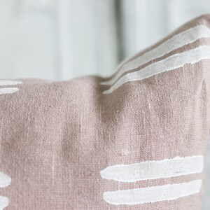 May include: Close-up of a dusty rose-colored pillow with white, horizontal brushstroke designs. The pillow has a textured linen appearance and a dark green tassel. This decorative pillow adds a touch of modern style to any room.