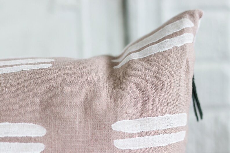 May include: Close-up of a dusty rose-colored pillow with white, horizontal brushstroke designs. The pillow has a textured linen appearance and a dark green tassel. This decorative pillow adds a touch of modern style to any room.