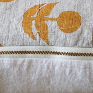 Puede incluir: Primer plano de un coj&iacute;n decorativo con fondo blanco y un estampado de dise&ntilde;os florales amarillo-naranja. Una cremallera de lat&oacute;n recorre horizontalmente el coj&iacute;n, separando la parte superior estampada de la inferior blanca lisa.