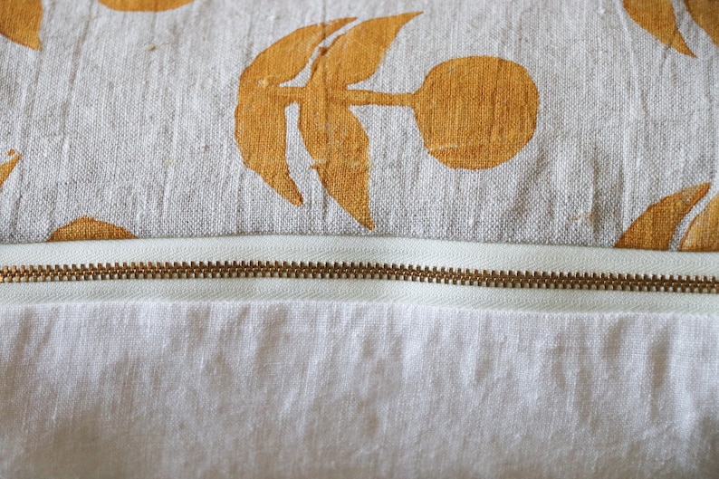 Puede incluir: Primer plano de un coj&iacute;n decorativo con fondo blanco y un estampado de dise&ntilde;os florales amarillo-naranja. Una cremallera de lat&oacute;n recorre horizontalmente el coj&iacute;n, separando la parte superior estampada de la inferior blanca lisa.