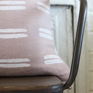 May include: A decorative pillow with a dusty rose background and white horizontal brushstroke patterns. The pillow is resting on a dark brown metal chair. The background includes a white brick wall and a white door.